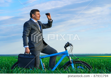 business concept - A businessman rides a bicycle on a green grass field, looks into the distance through binoculars, dressed in a business suit, he has a briefcase and documents 87931794