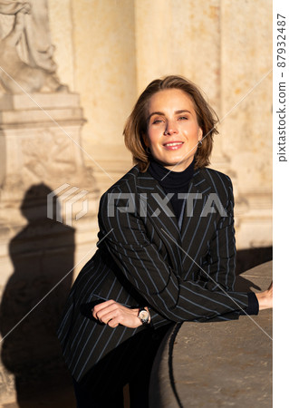 Young blond happy woman in striped jacket leaned on sunny historic building Young blond happy woman in striped jacket leaned on sunny historic building 87932487