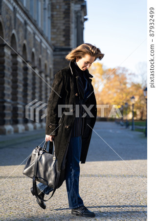 Young blond short-haired woman in long coat, jeans with bag walk on sunny street Young blond short-haired woman in long coat, jeans with bag walk on sunny street 87932499