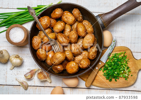 fried potatoes in a frying pan, garlic and green onions, farm eggs and salt, raw and cooked food in a rustic style on a background of white wooden boards 87933598