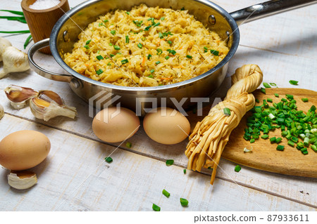 fried chopped cabbage in a frying pan, garlic and green onions, cheese, farm eggs and salt, raw and cooked food in a rustic style on a background of white wooden boards 87933611