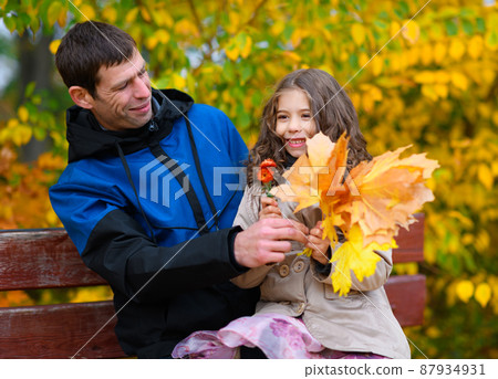 Father and daughter portrait in an autumn park. Happy people pose against the background of beautiful yellow trees. They sit on a bench, hug and are happy together. 87934931