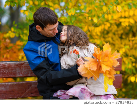 Father and daughter portrait in an autumn park. Happy people pose against the background of beautiful yellow trees. They sit on a bench, hug and are happy together. 87934933
