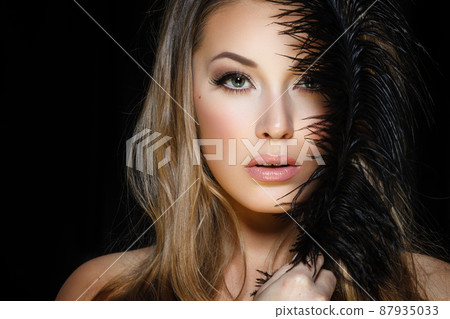Perfect beauty and glamour concept. Portrait of beautiful female model holding black feather on dark background. Young blond woman shows glamorous finery 87935033