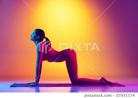 Portrait of young fit girl doing yoga back stretching exercises isolated over gradient pink and yellow background in neon 87935574