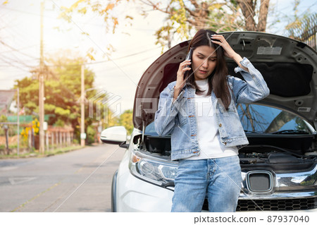 Angry Asian woman and using mobile phone calling for assistance after a car breakdown on street. Concept of vehicle engine problem or accident and emergency help from Professional mechanic 87937040