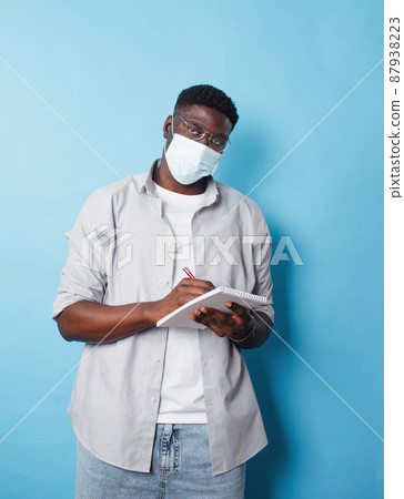 An African-American man in a medical mask writes information in a notebook on an isolated blue background 87938223