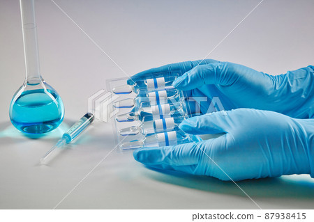 Hands in disposable gloves holding set of ampoules with liquid. Medical flask with blue chemical reagent and syringe isolated on white. Coronavirus 87938415