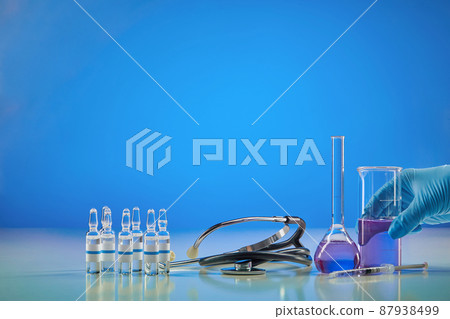 Coronavirus. Hand in glove holding beaker with purple reagent. Six ampoules with liquid, syringe, medical flask and phonendoscope, blue background. 87938499
