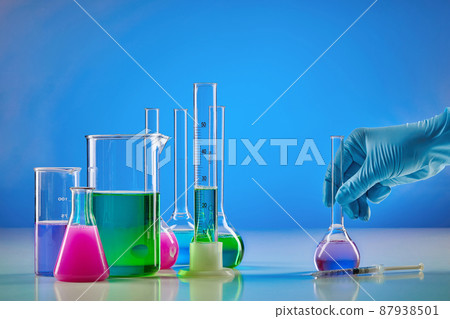 Hand in disposable glove holding medical flask. Colorful chemical reagents in beakers and measuring cylinder, blue background. Coronavirus pandemic 87938501