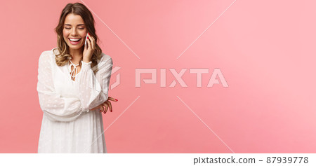 Communication and technology concept. Attractive caucasian female in white dress, blond short haircut, hold smartphone, talking on mobile phone and laughing, smiling carefree, pink background 87939778