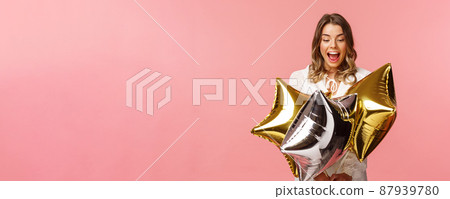 Holidays, celebration and women concept. Portrait of joyful beautiful blond girl in white dress being excited and impressed with cool start-shaped balloons, partying over pink background Holidays, celebration and women concept. Portrait of joyful beautiful blond girl in white dress being excited and impressed with cool start-shaped balloons, partying over pink background 87939780