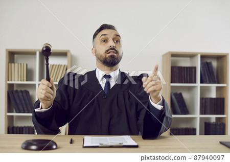 Judge in court of law considers the case closed and passes judgement. Young man in a robe uniform sitting at a table in the courtroom pronounces a fair sentence and hits the sound block with a gavel Judge in court of law considers the case closed and passes judgement. Young man in a robe uniform sitting at a table in the courtroom pronounces a fair sentence and hits the sound block with a gavel 87940097