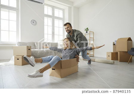 Young married couple having fun on moving day. Happy first time buyers playing with boxes in modern spacious living room interior in new home. Real estate, mortgage, buying house of your dream concept 87940135