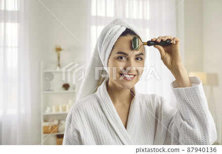 Portrait of happy young woman after shower massaging face with trendy smooth facial beauty roller to stimulate lymphatic system, relax muscles, reduce puffiness and increase blood flow for skin's glow 87940136