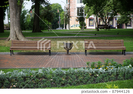Two empty wooden benches on the city alley. Waste bin between two benches. Two empty wooden benches on the city alley. Waste bin between two benches. 87944126