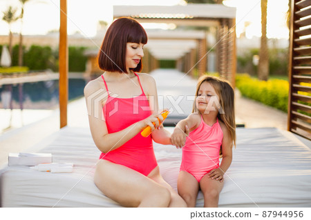 Young pretty woman applying sunscreen spray on hand of her daughter in a pink swimsuits 87944956