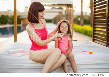 An adult woman in pink swimsuit with same dressed daughter and dabbed her with wipe for cleaning 87944976
