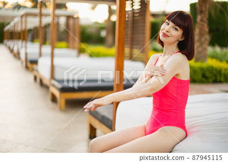 Beautiful caucasian woman in a pink swimsuit applying putting skin care sunscreen lotion. 87945115