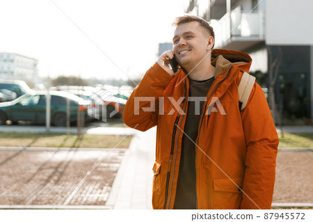 Handsome young man using his phone with smile, guy standing on the street in the city. So glad to hear you. Portrait of excited happy man talking with friend on phone. Urban lifestyle concept. Handsome young man using his phone with smile, guy standing on the street in the city. So glad to hear you. Portrait of excited happy man talking with friend on phone. Urban lifestyle concept. 87945572