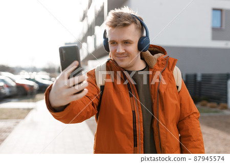 Young happy guy is making selfie on a phone, man walking in spring city outdoors. Urban lifestyle concept. Traveler. 87945574