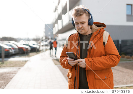 A handsome young man using his phone with headphones and standing on the street in the city. Music lover enjoying music. Urban lifestyle concept. Traveler. A handsome young man using his phone with headphones and standing on the street in the city. Music lover enjoying music. Urban lifestyle concept. Traveler. 87945576