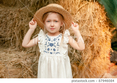 Cute little blonde girl in light dress with embroidery and straw hat standing on hayloft Cute little blonde girl in light dress with embroidery and straw hat standing on hayloft 87946084