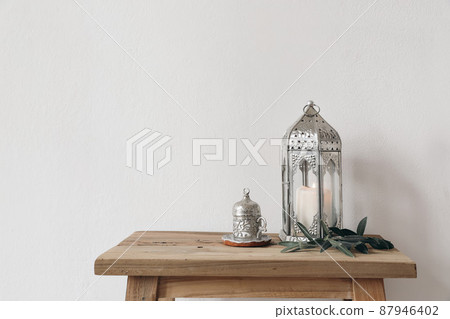 Ramadan Kareem still life. Ornamental lantern with burning candle and Turkish silver cup with tea or coffee. Green olive tree branches on old wooden table background. Muslim Iftar dinner. No people 87946402