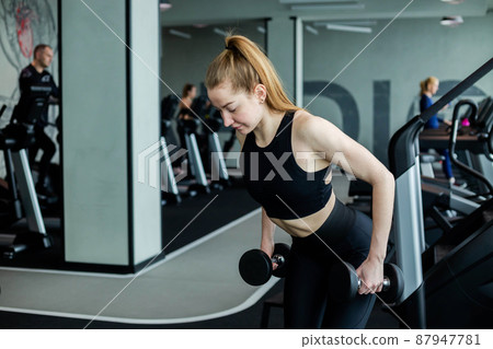 A young girl in black sportswear is engaged in the gym with dumbbells. A young girl in black sportswear is engaged in the gym with dumbbells. 87947781