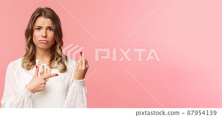 Close-up portrait sad and frustrated young blond girl waited for proposal during romantic date, pointing at finger without weddint ring with puzzled upset expression, standing pink background 87954139