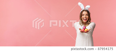 Holidays, spring and party concept. Portrait of lovely, romantic young blond woman in rabbit ears and white dress, giving you painted easter eggs as celebrating orthodox holiday, pink background 87954175