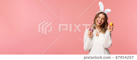 Holidays, spring and party concept. Cheerful good-looking blond woman celebrating Easter day in rabbit ears, holding two painted eggs and wink camera, smiling happily, pink background Holidays, spring and party concept. Cheerful good-looking blond woman celebrating Easter day in rabbit ears, holding two painted eggs and wink camera, smiling happily, pink background 87954207
