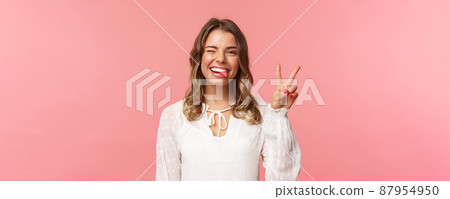 Close-up portrait of friendly joyful blond girl in white dress, sending positive vibes, smiling pleased and wink, stick tongue showing peace sign, looking kawaii over pink background 87954950