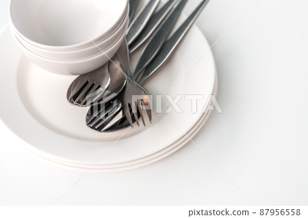 White food dishes, small bowls, forks, and spoons together, stacks dishes, bowl and group of fork and spoons on white table, natural light, view from above, space for text. 87956558