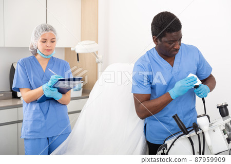 Female cosmetologist filling medical form, male assistant preparing equipment 87959909