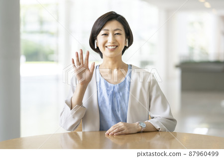 A smiling business woman in her thirties wearing wireless earphones waving at the camera 87960499