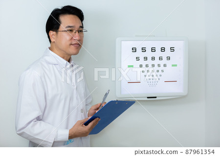 Man ophthalmologist wear uniform holding clipboard standing near eye chart vision test in clinic. 87961354