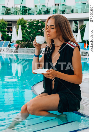 Young blonde woman sitting next to swimming pool drinking coffee in hotel on vacation. Blogger on holidays. 87961694