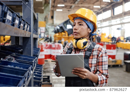Young female constructor with tablet standing by shelf with containers Young female constructor with tablet standing by shelf with containers 87962627
