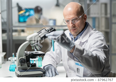 Contemporary mature male clinician looking at blue liquid in flask Contemporary mature male clinician looking at blue liquid in flask 87962767