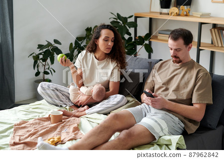 Young woman looking at screen of smartphone of her husband sitting next to her Young woman looking at screen of smartphone of her husband sitting next to her 87962842