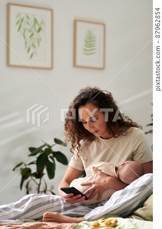 Young mother with baby on hands texting in mobile phone while sitting on bed Young mother with baby on hands texting in mobile phone while sitting on bed 87962854