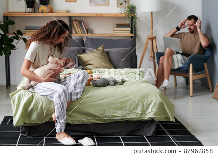 Young female putting newborn baby to sleep while sitting on double bed Young female putting newborn baby to sleep while sitting on double bed 87962858