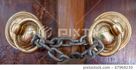 Closeup of two antique copper ornate door knockers over an aged wooden ornate door closed with rusted chain Closeup of two antique copper ornate door knockers over an aged wooden ornate door closed with rusted chain 87969230
