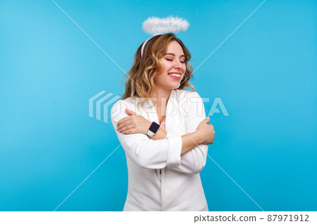 Portrait of egoistic beautiful angelic woman with halo on her wavy hair and in white clothes embracing herself, standing with eyes closed from pleasure and happiness. studio shot, blue background Portrait of egoistic beautiful angelic woman with halo on her wavy hair and in white clothes embracing herself, standing with eyes closed from pleasure and happiness. studio shot, blue background 87971912