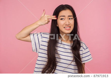 Suicide gesture. Portrait of depressed girl with brunette hair in t-shirt aiming finger gun to head with helpless desperate expression, shooting herself. indoor studio shot isolated on pink background Suicide gesture. Portrait of depressed girl with brunette hair in t-shirt aiming finger gun to head with helpless desperate expression, shooting herself. indoor studio shot isolated on pink background 87971913