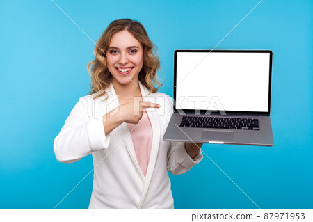Look at internet advertisement. Portrait of happy businesswoman with wavy hair in white jacket pointing laptop with blank screen and smiling at camera. indoor studio shot isolated on blue background Look at internet advertisement. Portrait of happy businesswoman with wavy hair in white jacket pointing laptop with blank screen and smiling at camera. indoor studio shot isolated on blue background 87971953