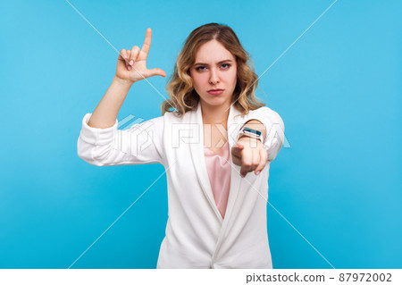 You are loser. Portrait of irritated woman with wavy hair in white jacket doing loser gesture, L finger sign and pointing at camera, blaming for failures. studio shot isolated on blue background You are loser. Portrait of irritated woman with wavy hair in white jacket doing loser gesture, L finger sign and pointing at camera, blaming for failures. studio shot isolated on blue background 87972002