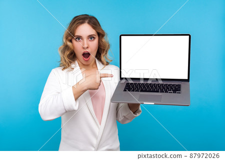 Amazing internet advertising. Portrait of shocked businesswoman with wavy hair in white jacket pointing laptop with blank screen, looking surprised about news. studio shot isolated on blue background Amazing internet advertising. Portrait of shocked businesswoman with wavy hair in white jacket pointing laptop with blank screen, looking surprised about news. studio shot isolated on blue background 87972026