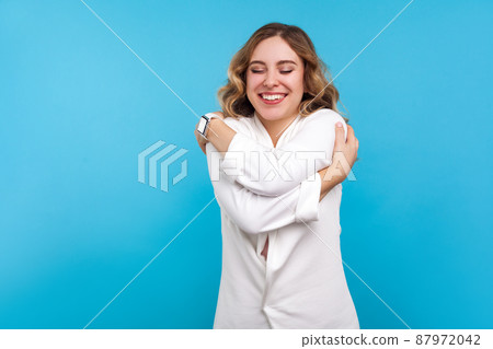 Self-esteem, love. Portrait of egoistic beautiful woman with wavy hair in white jacket embracing herself, standing with eyes closed from pleasure and happiness. studio shot isolated on blue background Self-esteem, love. Portrait of egoistic beautiful woman with wavy hair in white jacket embracing herself, standing with eyes closed from pleasure and happiness. studio shot isolated on blue background 87972042